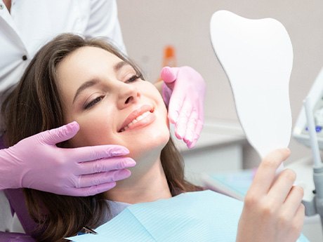 Woman in dental chair smiling at reflection with dentist in pink gloves touching her face