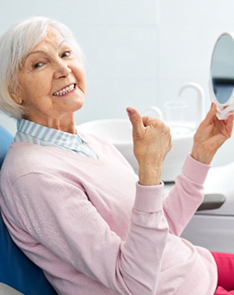 Senior woman holding mirror and giving thumbs up in dental chair